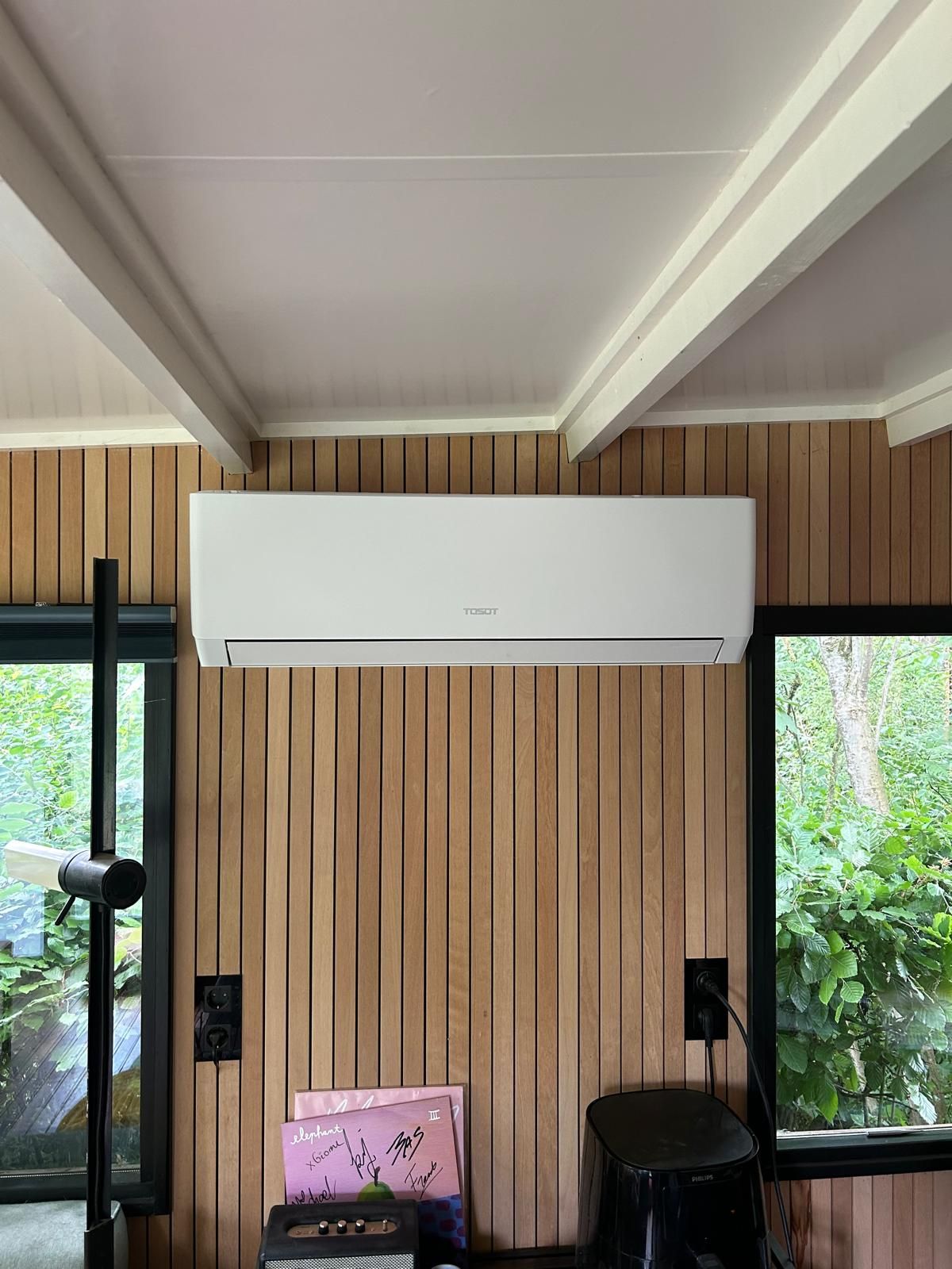 Witte airconditioningunit gemonteerd aan een houten wand tussen de ramen; een zwart onderstel is zichtbaar.