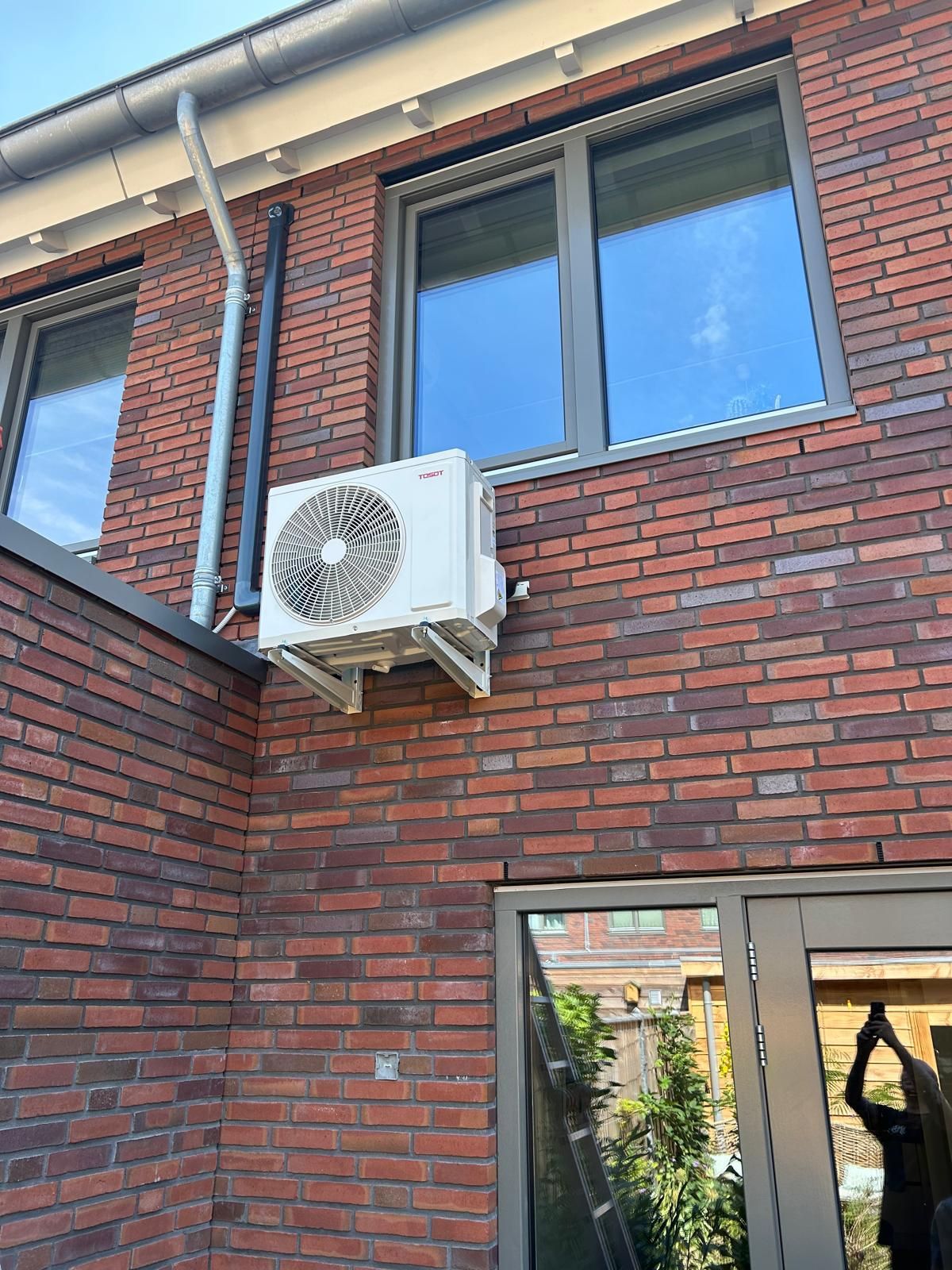 Airconditioningunit gemonteerd aan de buitenkant van een bakstenen gebouw, vlakbij een raam en een dakgoot.