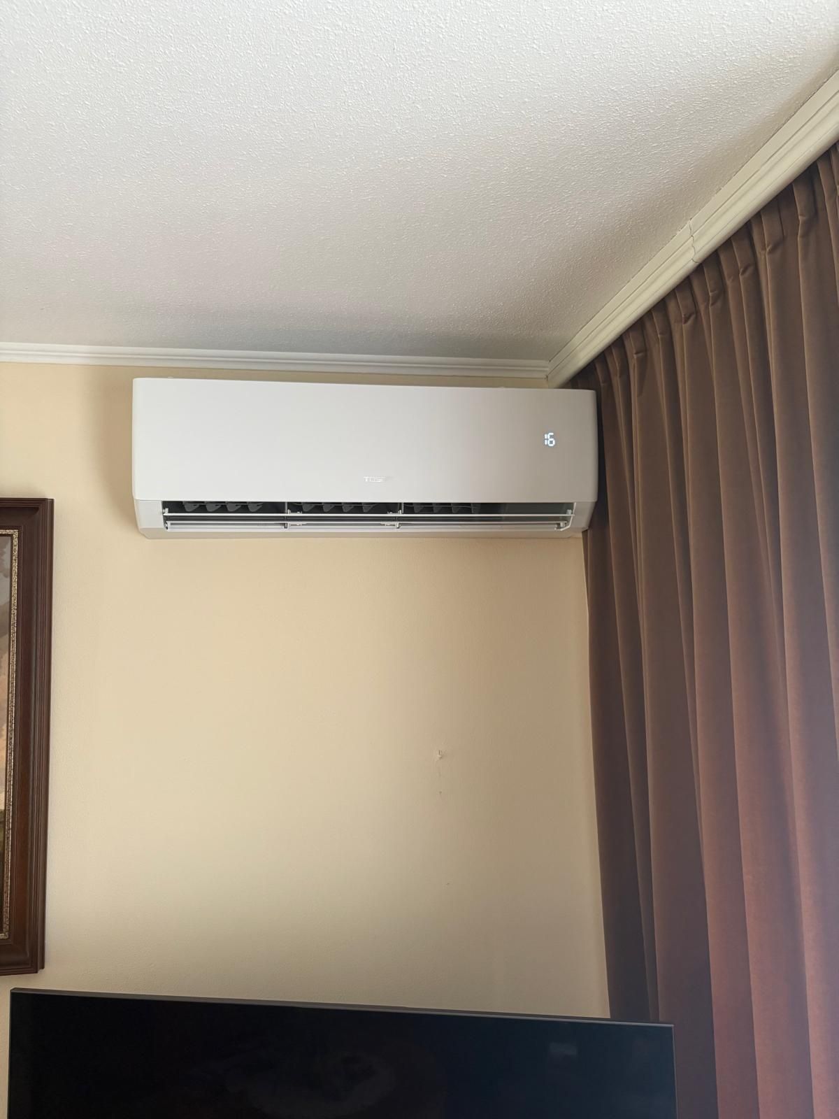 Witte, aan de muur gemonteerde airconditioner op een beige muur naast een bruin gordijn.