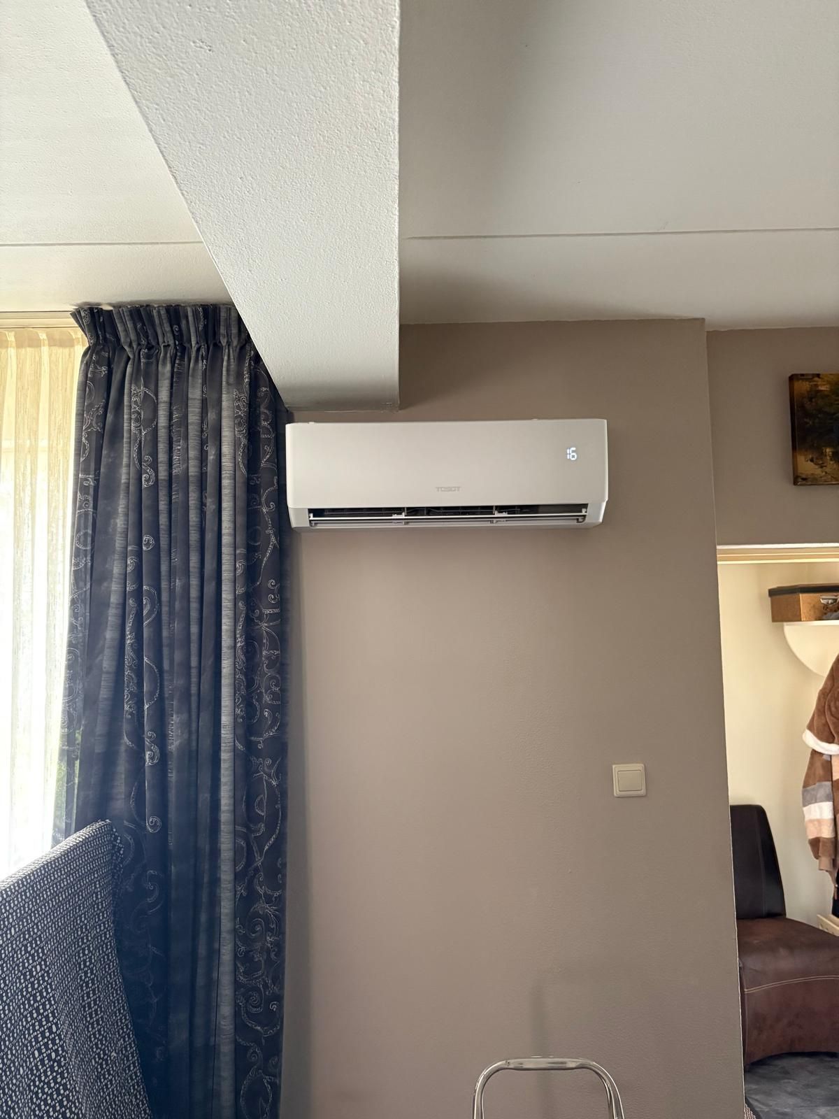 Airconditioningunit gemonteerd aan een taupekleurige muur, met een donker gordijn aan de linkerkant en een deuropening aan de rechterkant.