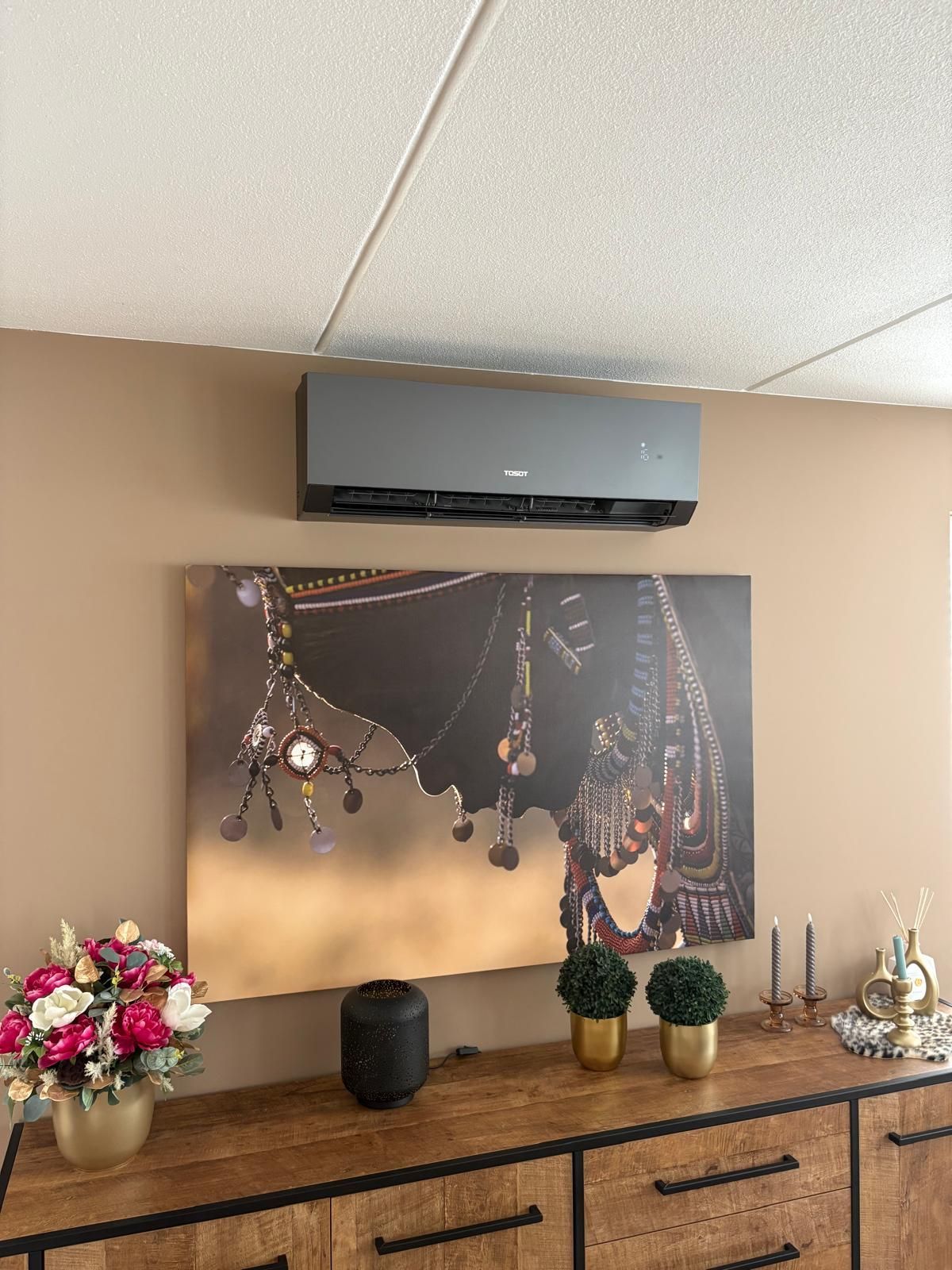 Een moderne airconditioningunit boven een consoletafel met decoratie, waaronder een schilderij en bloemen, binnenshuis.