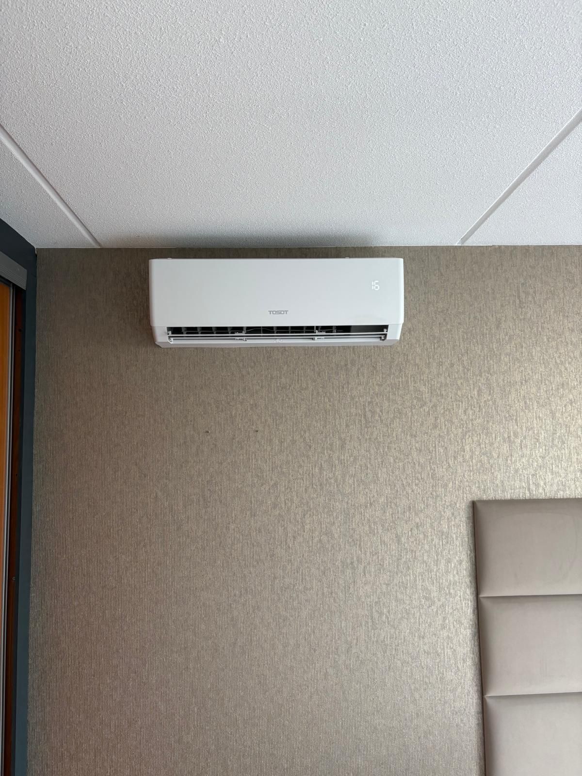 Witte airconditioner gemonteerd aan een lichtbruine muur met structuurverf. Een deel van het witte plafond bevindt zich erboven.