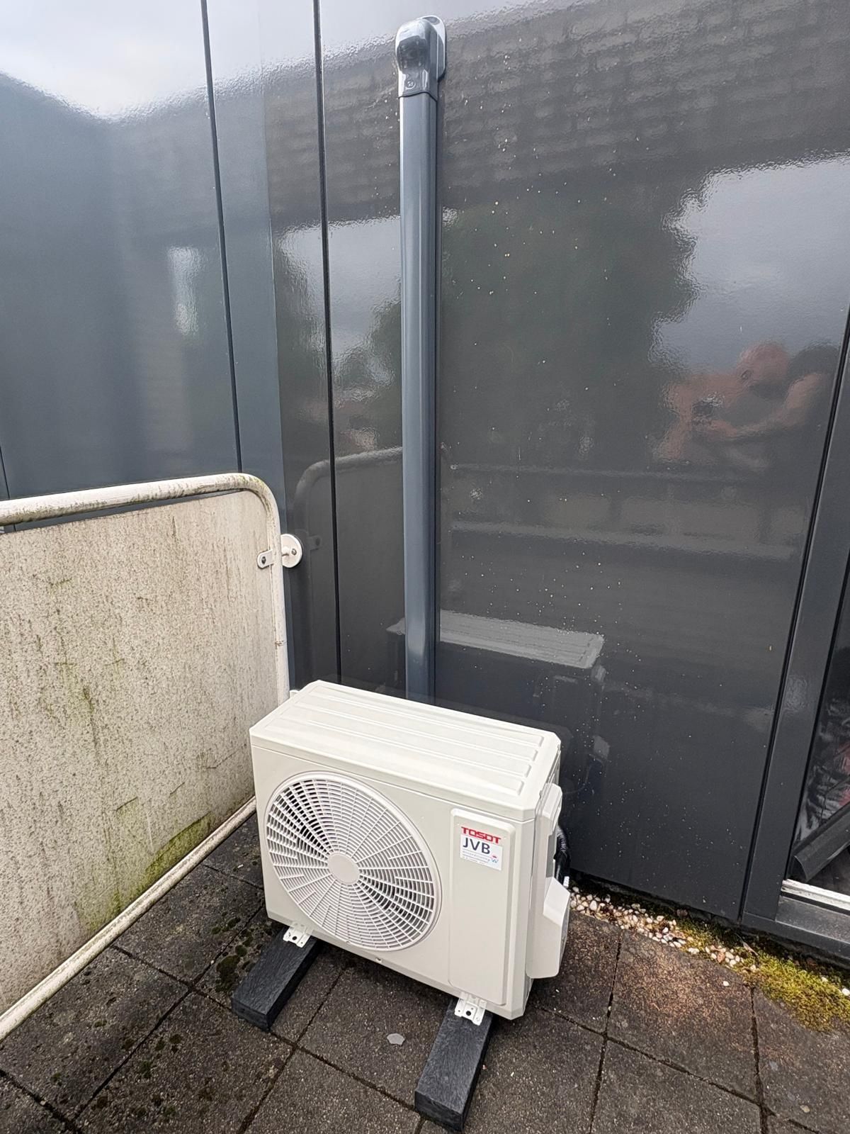 Een lichtgekleurde buitenunit van de airconditioning, op een zwarte standaard, tegen een donkergrijze muur, vlakbij een regenpijp.