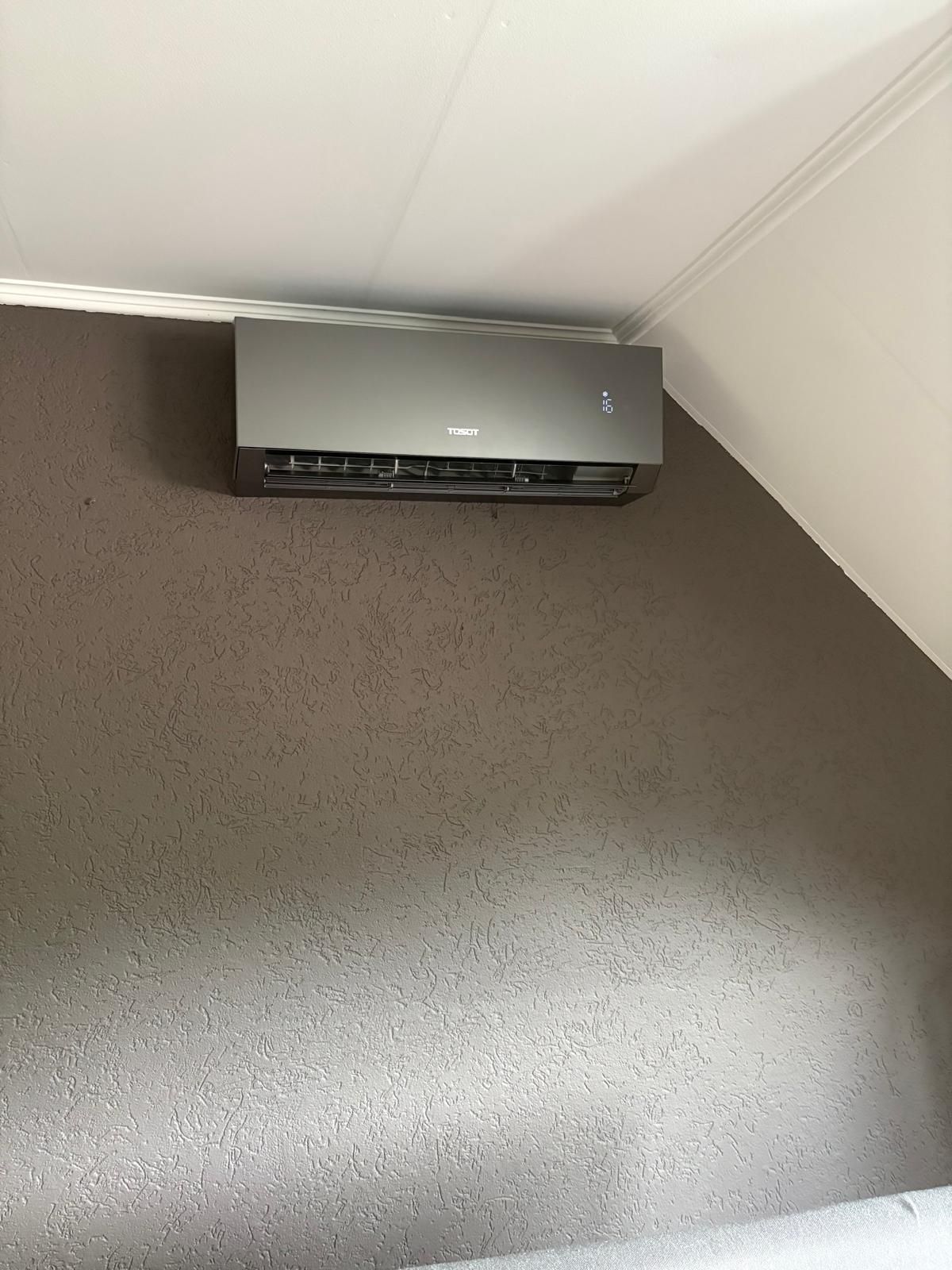 Zilverkleurige airconditioningunit gemonteerd op een grijze, gestructureerde muur. Wit plafond en een schuine muurhoek zichtbaar.