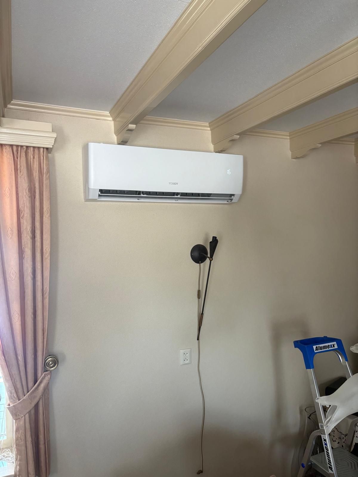 Witte wandairconditioner op een beige muur, met hangend snoer en gordijnen.