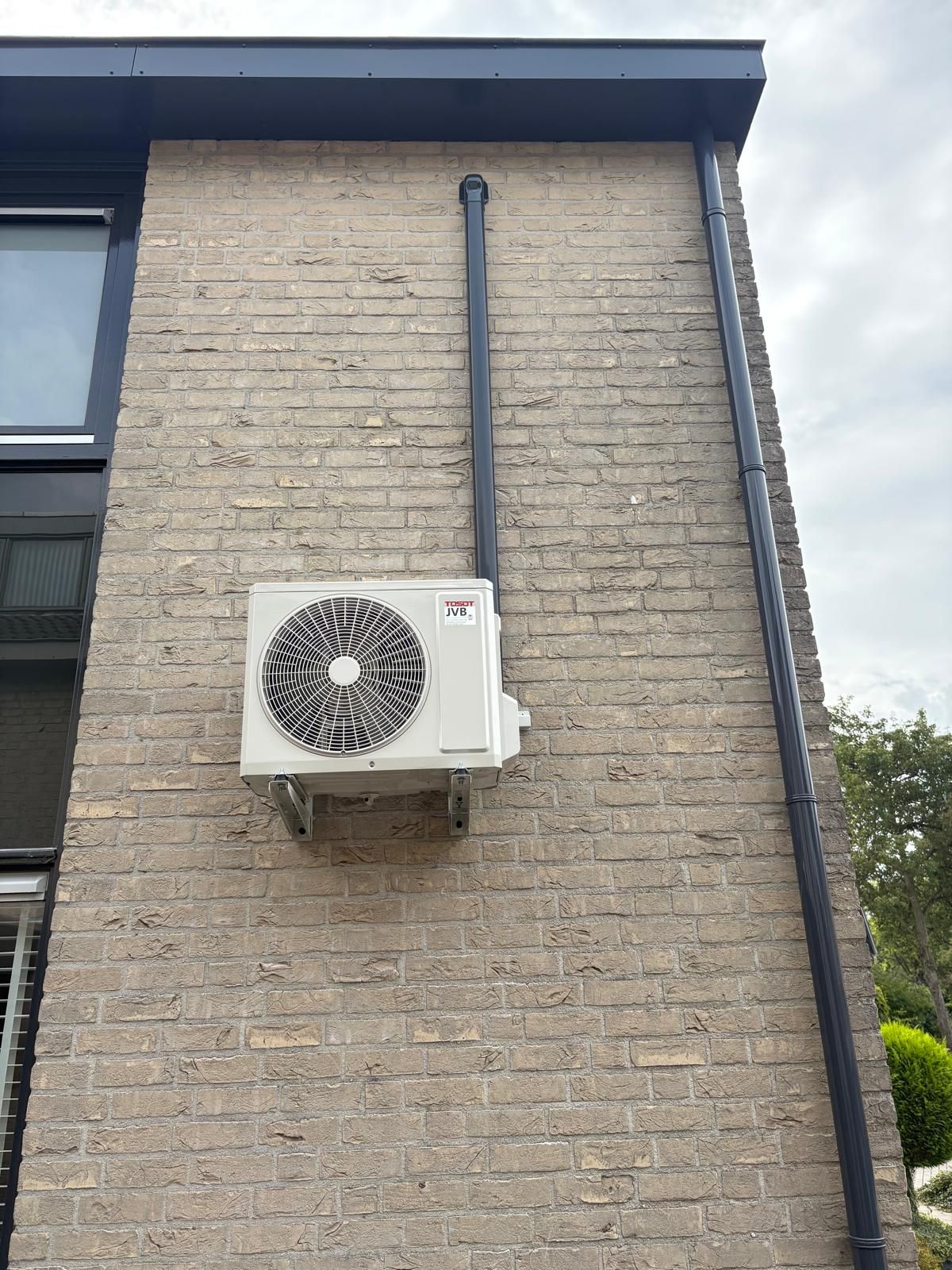 Een airconditioningunit gemonteerd op een bakstenen buitenmuur met zwarte leidingen.