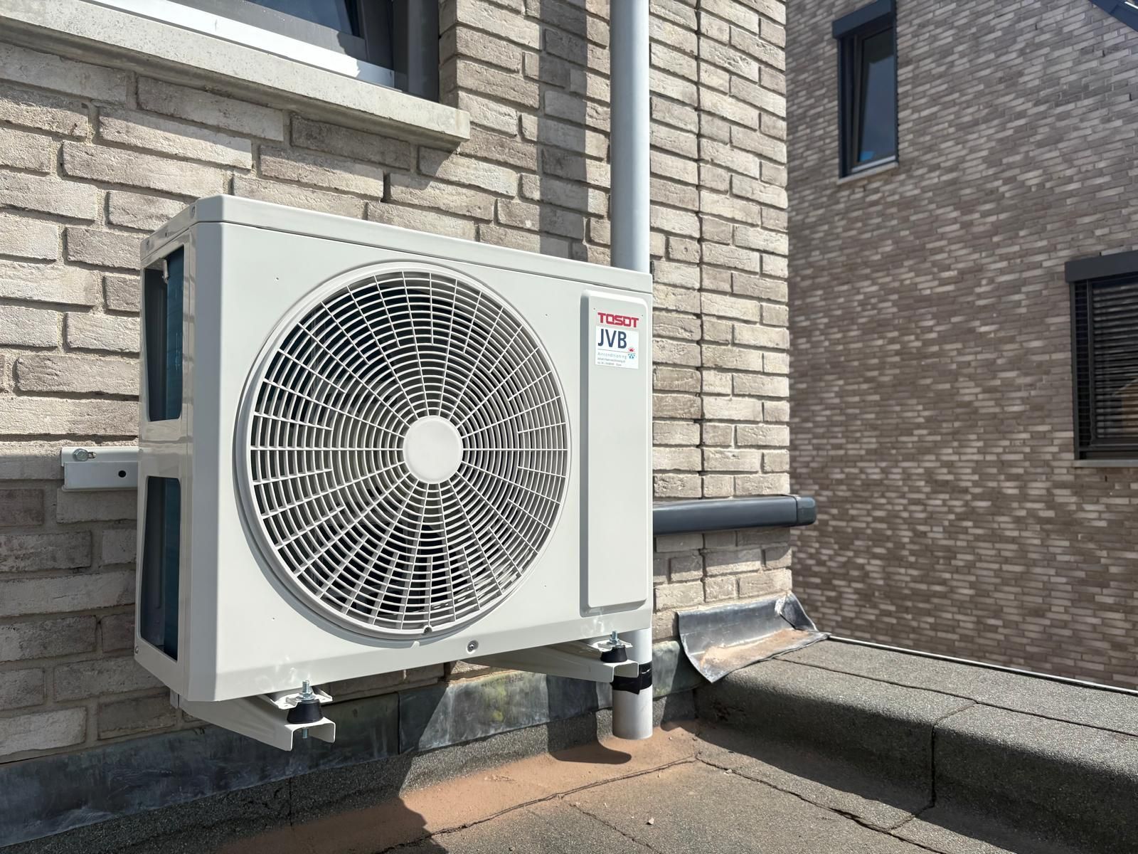 Buitenunit voor airconditioning, gemonteerd op een bakstenen muur, met ventilator en leidingen.