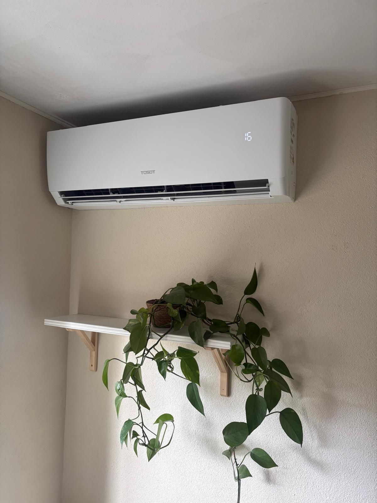 Witte airconditioner boven een plank met een potplant aan een beige muur.