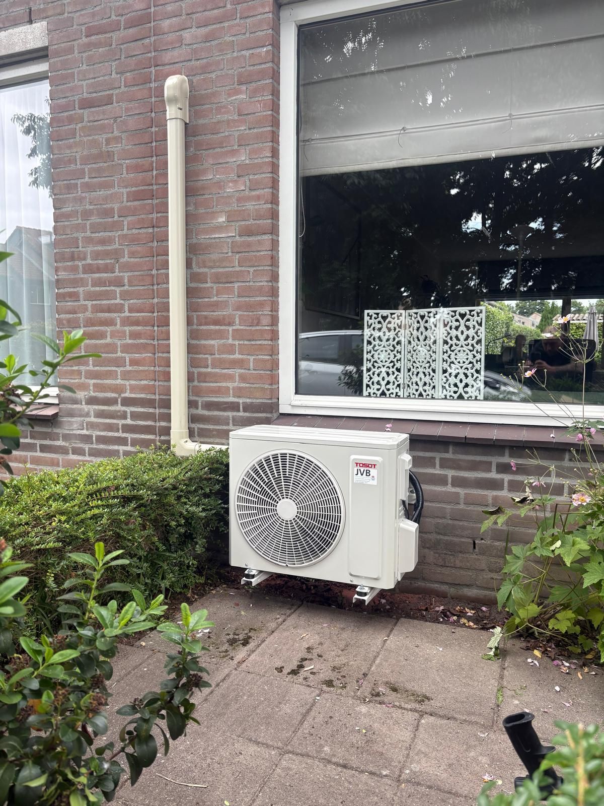 Airconditioningunit buiten een bakstenen gebouw, naast een raam en struiken.