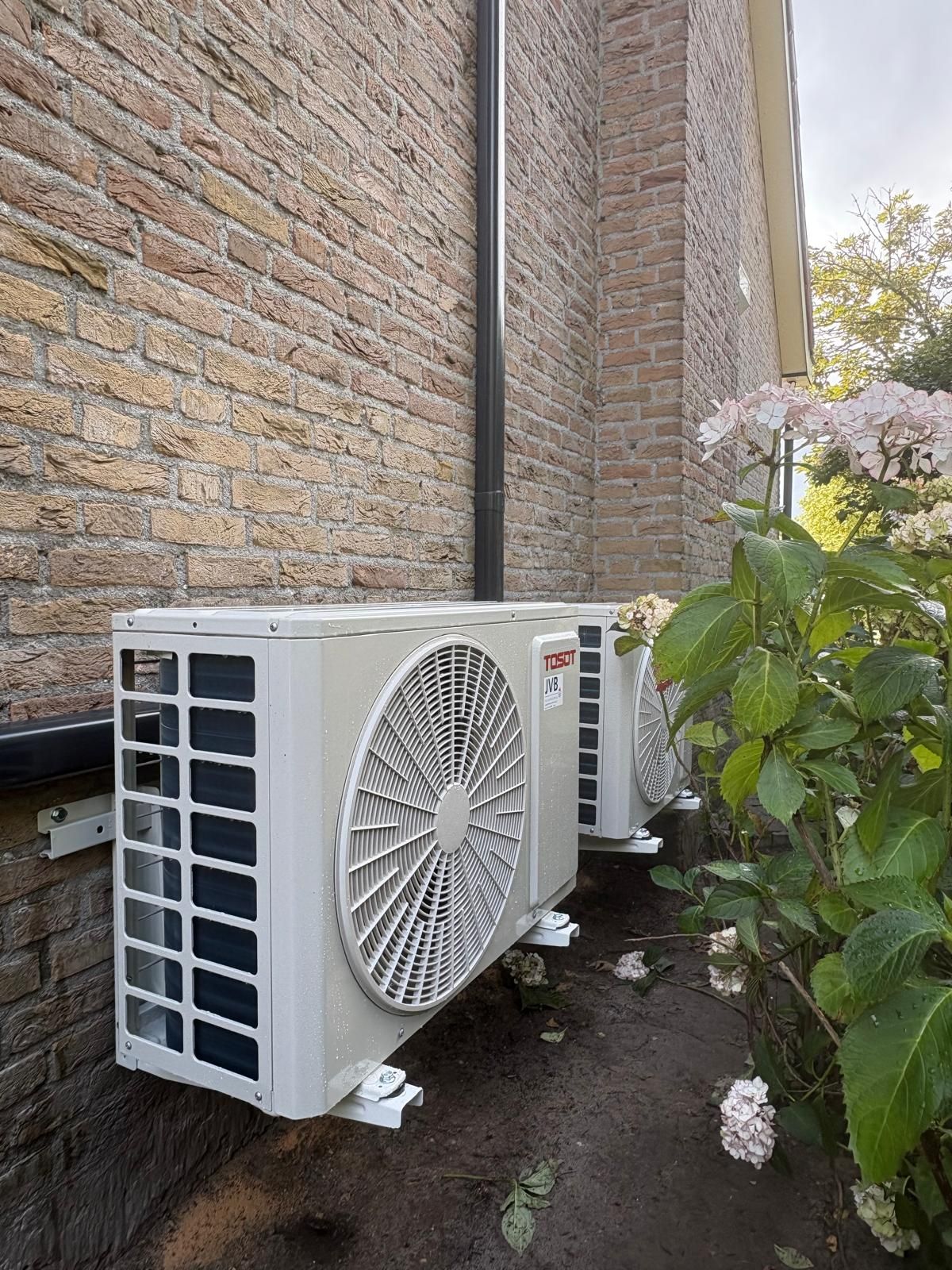 Drie rechthoekige airconditioningunits gemonteerd op een bakstenen muur naast een struik.