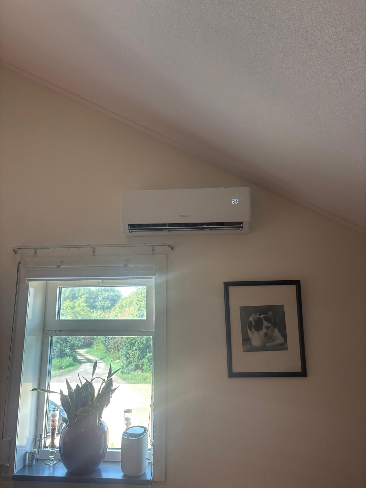 Witte airconditioningunit boven een raam, ingelijste kunst en een plant. Beige muren.