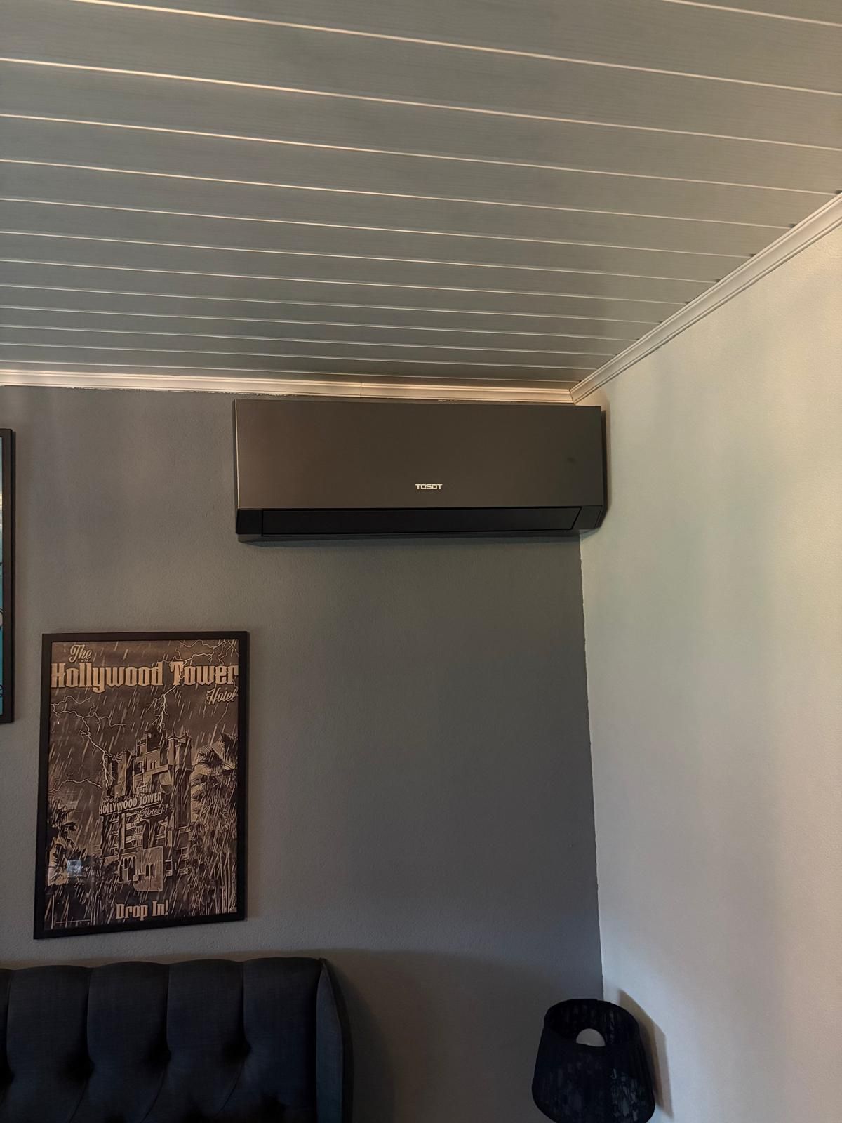 Zwarte airconditioner gemonteerd aan een donkergrijze muur, boven een bank en onder een plafond met structuurafwerking.