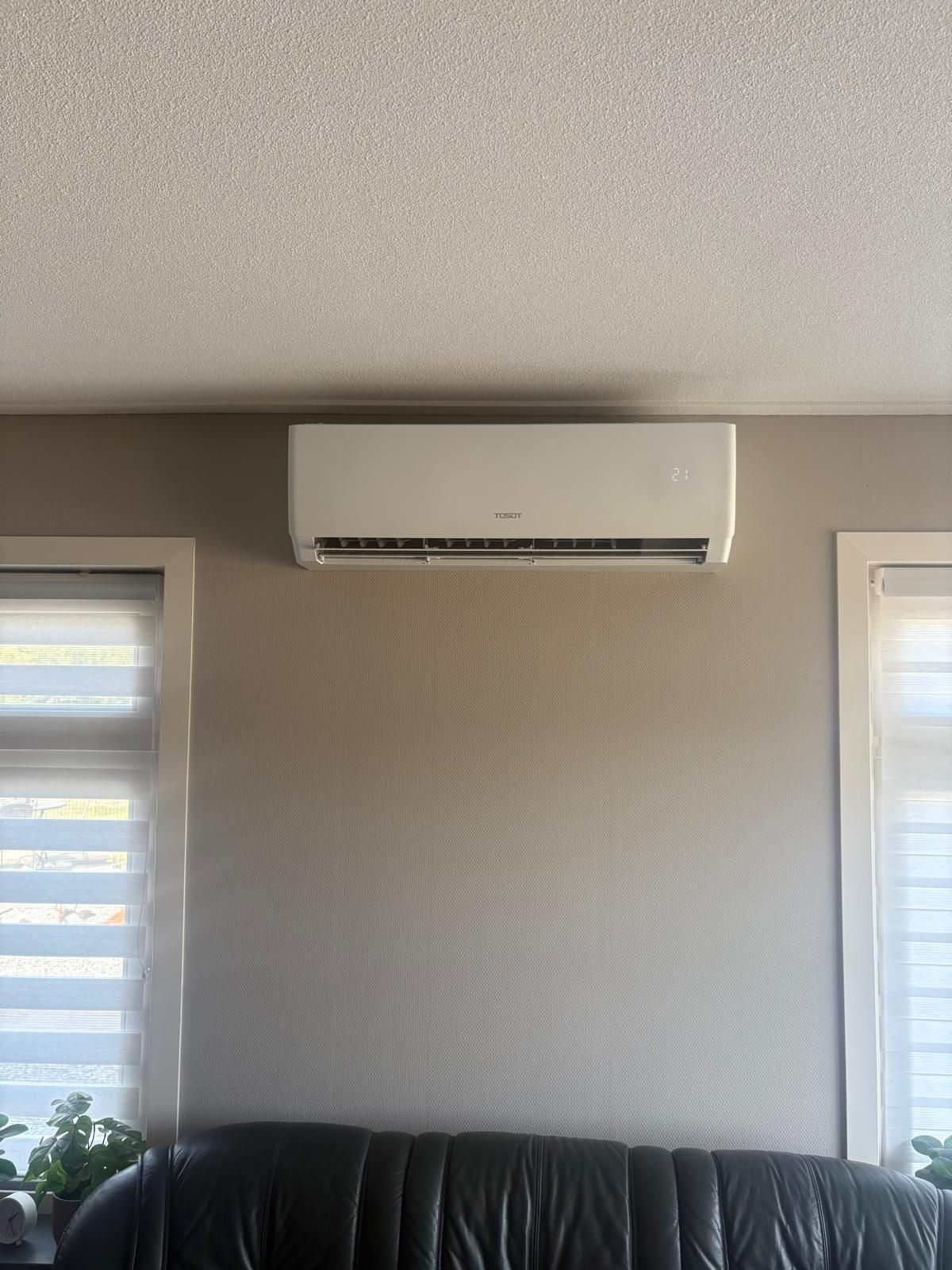 Een aan de muur gemonteerde airconditioner boven een zwarte bank, tussen twee ramen met jaloezieën, op een beige muur met structuur.