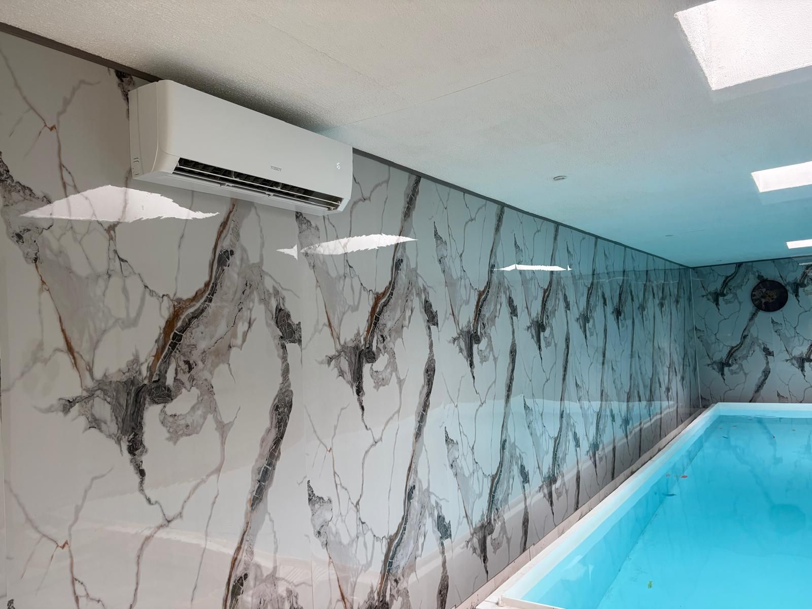 Een witte en grijze muur met marmerlook, met daarboven een airconditioningunit boven een binnenzwembad.