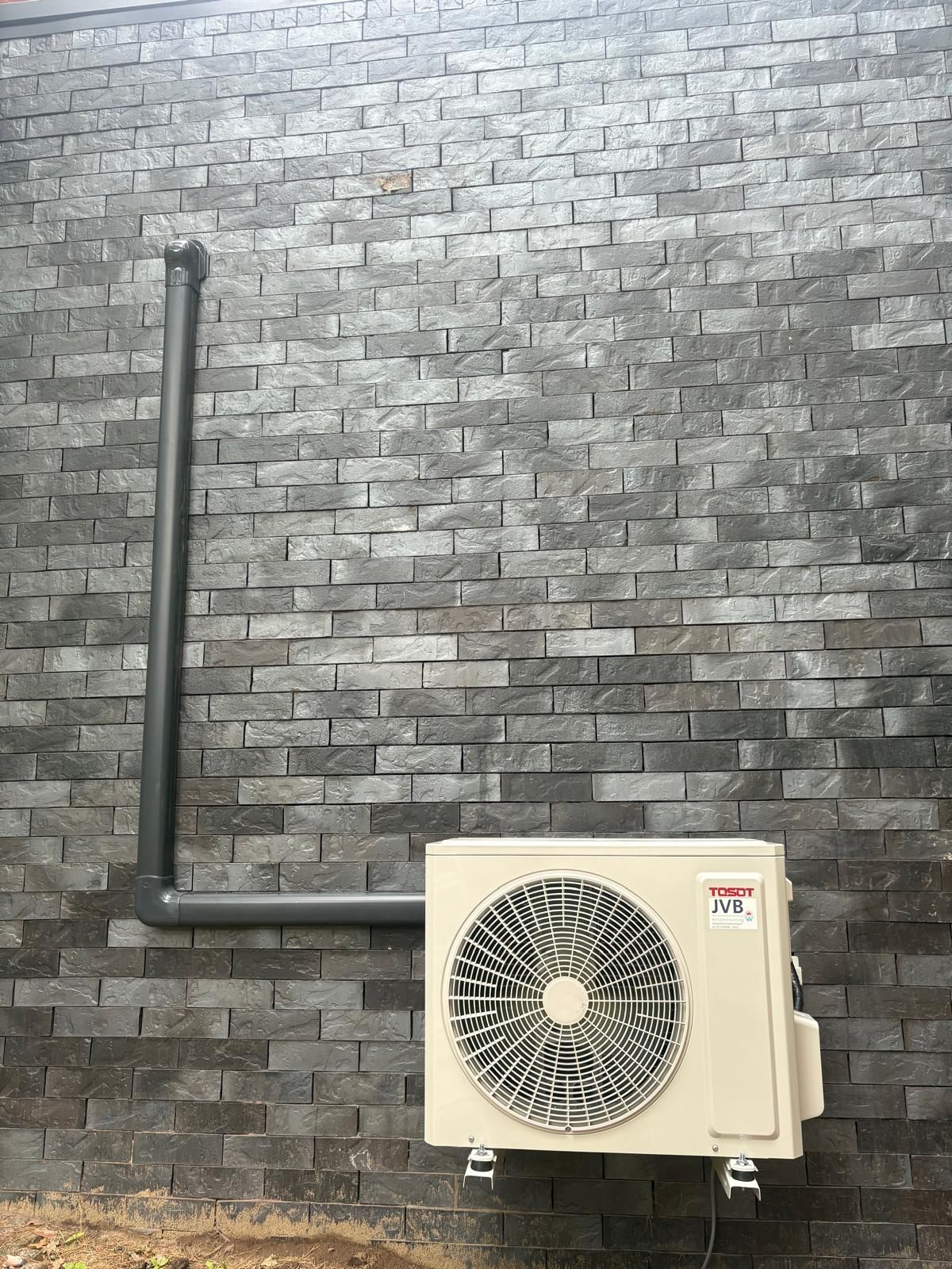 Airconditioningunit gemonteerd op een grijze bakstenen muur met donkergrijze afvoerpijp.