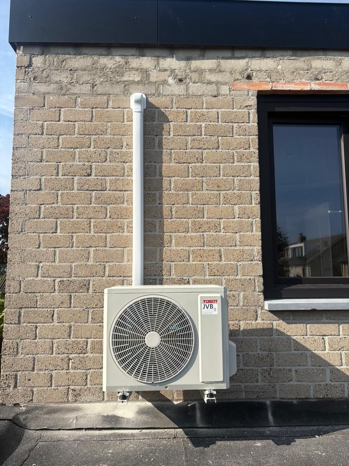 Airconditioningunit gemonteerd aan een bakstenen muur met witte afvoerpijp.