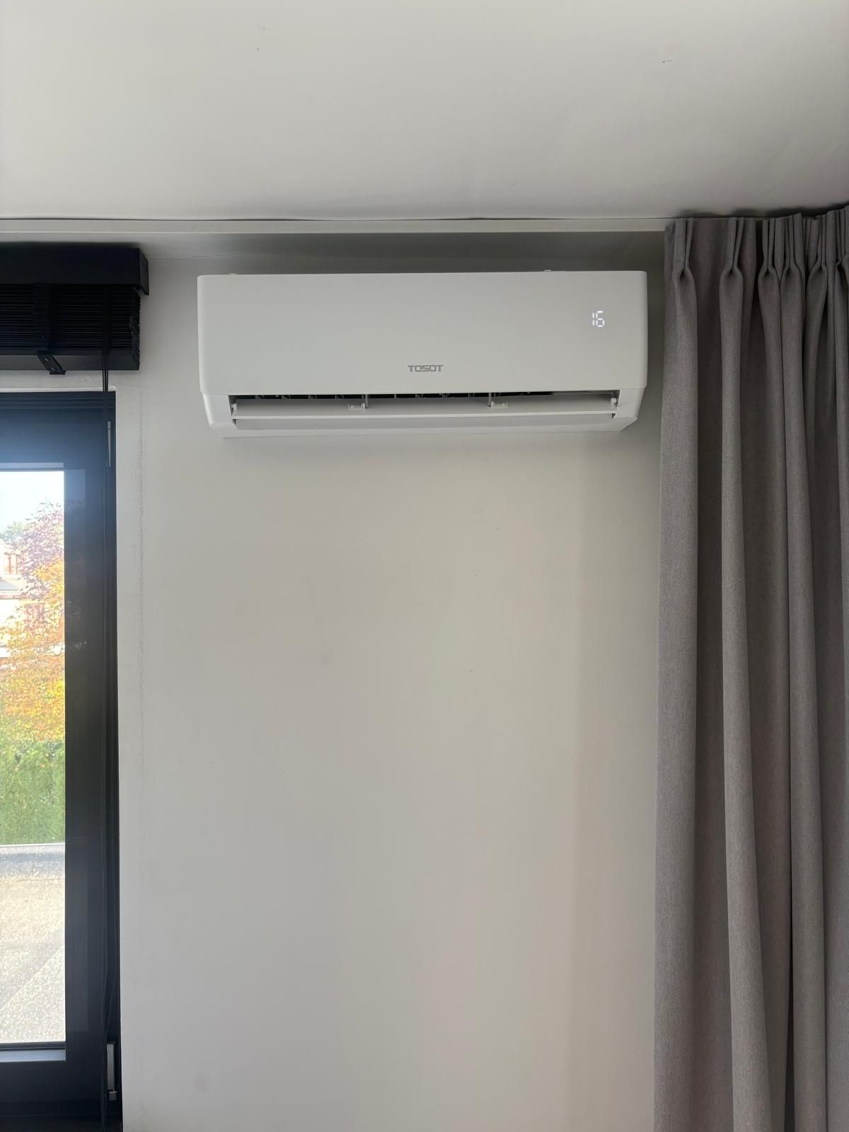 Witte, aan de muur gemonteerde airconditioner boven het raam. Rechts hangt een grijs gordijn.