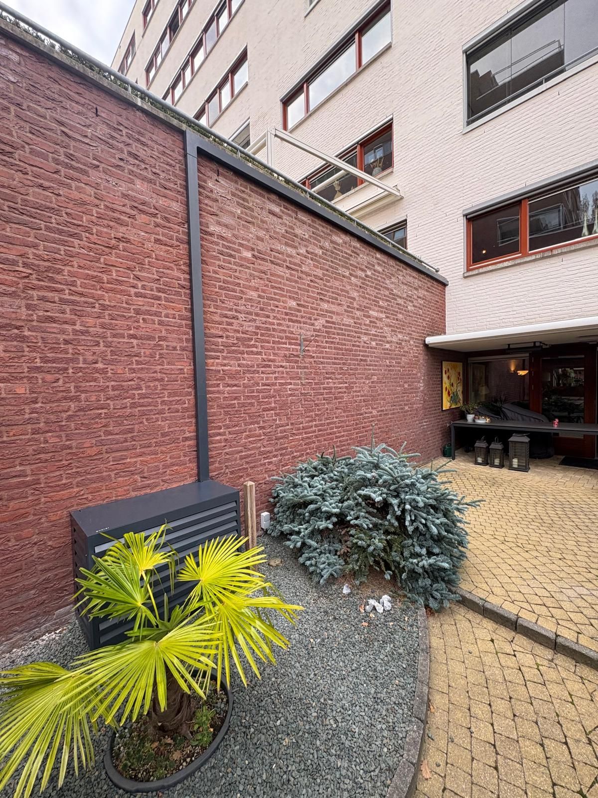 Een bakstenen muur, een buitenruimte met groen en een gebouw op de achtergrond.