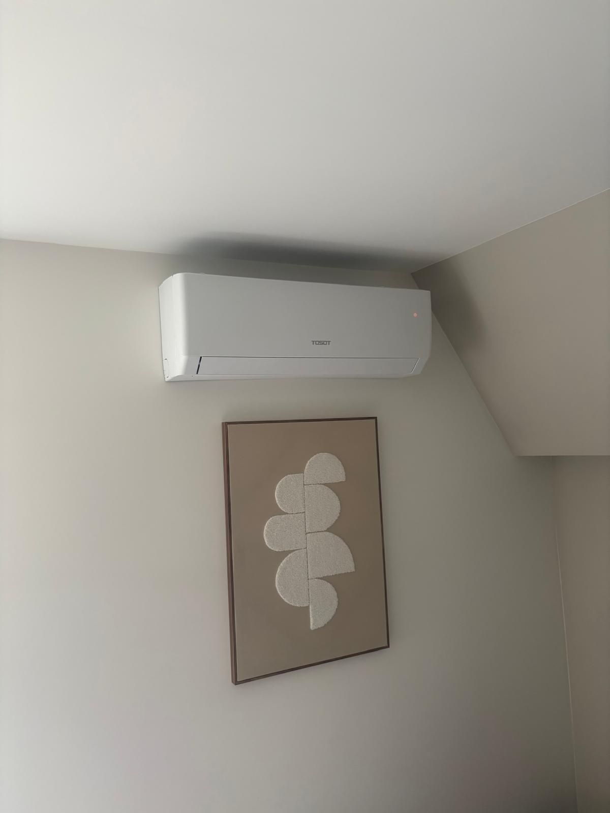 Witte airconditioningunit gemonteerd aan een lichte muur boven een ingelijst abstract kunstwerk.
