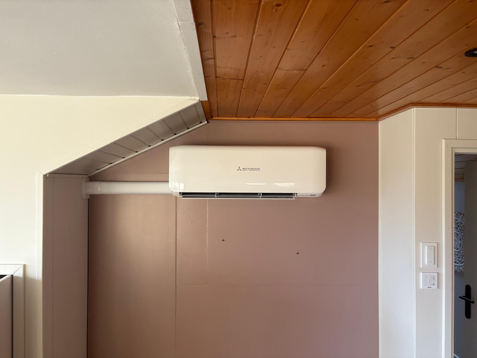 Airconditioning gemonteerd aan een roze muur onder een houten plafond.