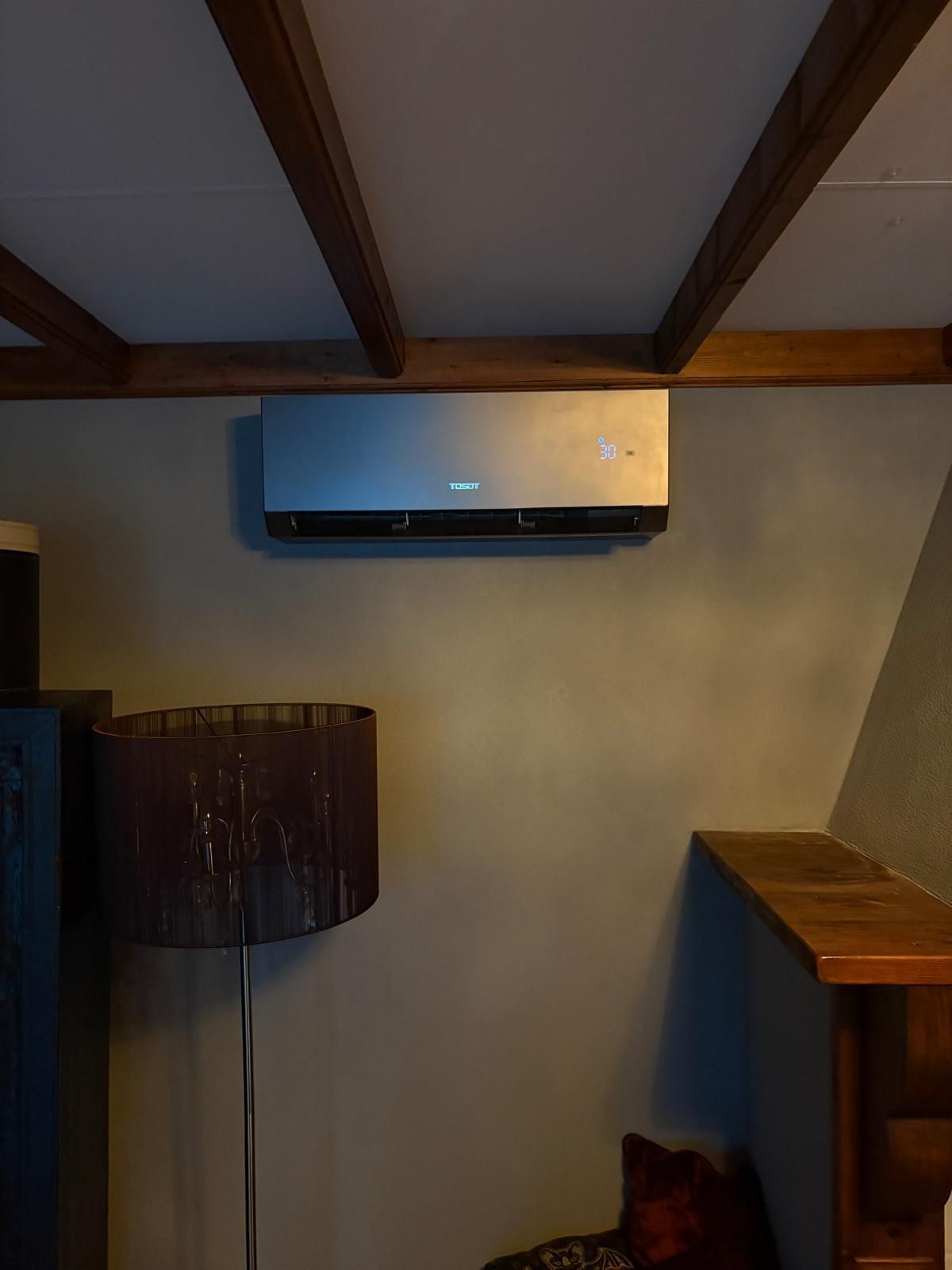 Airconditioningunit gemonteerd aan een beige muur, onder donkere houten balken. Een lamp en een hoek van een open haard zijn zichtbaar.
