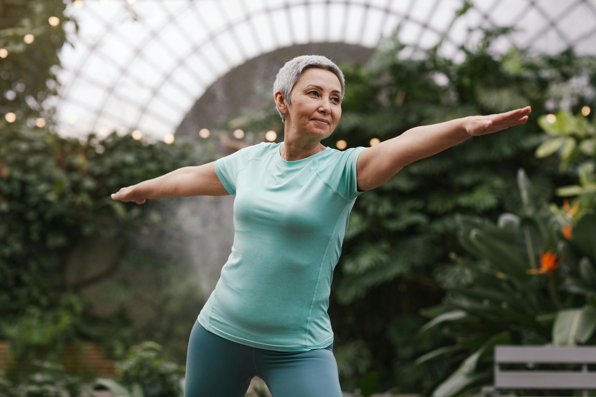 An older woman is practicing yoga in a greenhouse.
