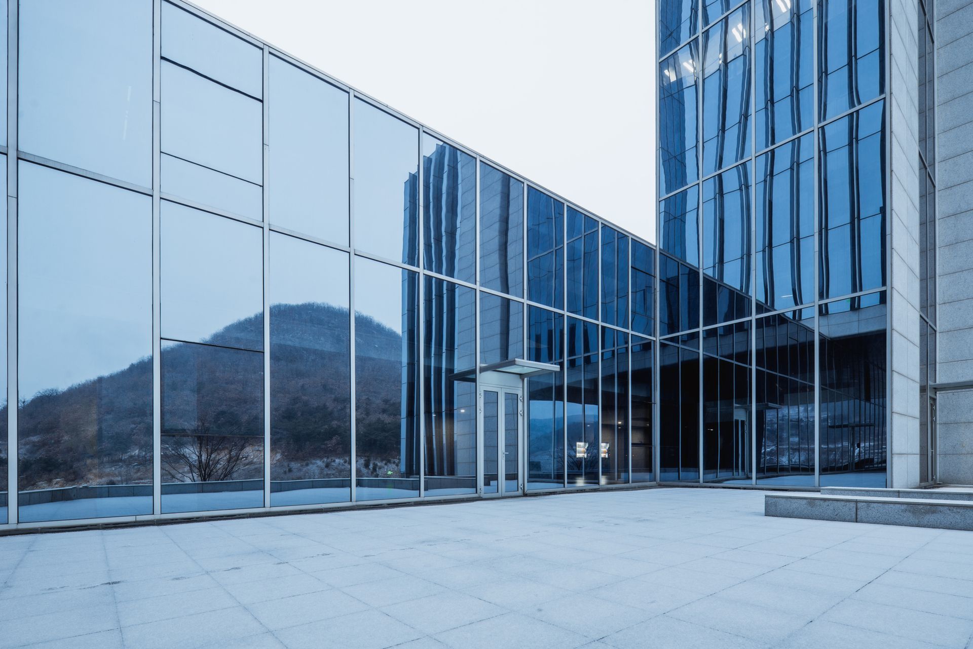 Modern glass building with a mountain reflected in the windows; overcast sky and stone patio.