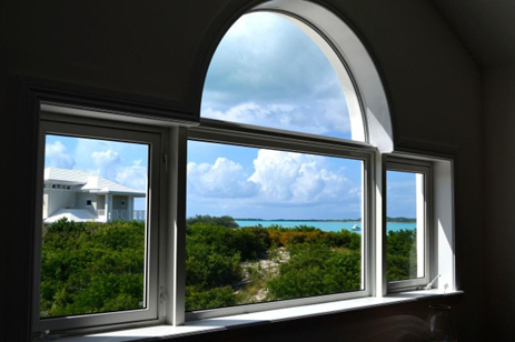 Large window overlooking a bright blue ocean and a white house on a green landscape under a cloudy sky.