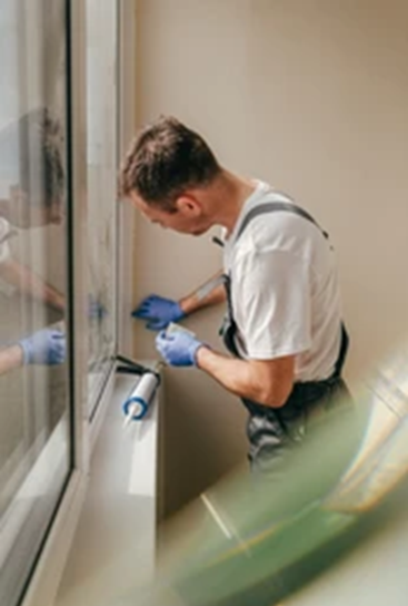Man in overalls caulking a window frame, wearing blue gloves, in a bright room.