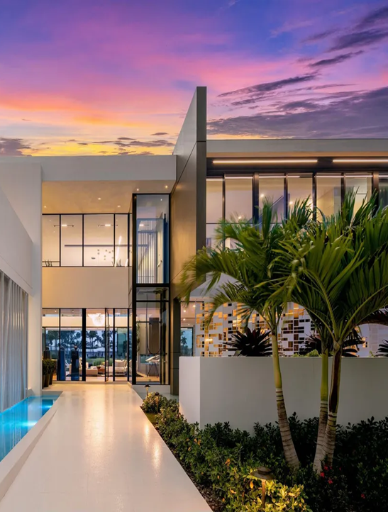 Modern white home with glass windows, palm trees, walkway, and pool against a sunset sky.
