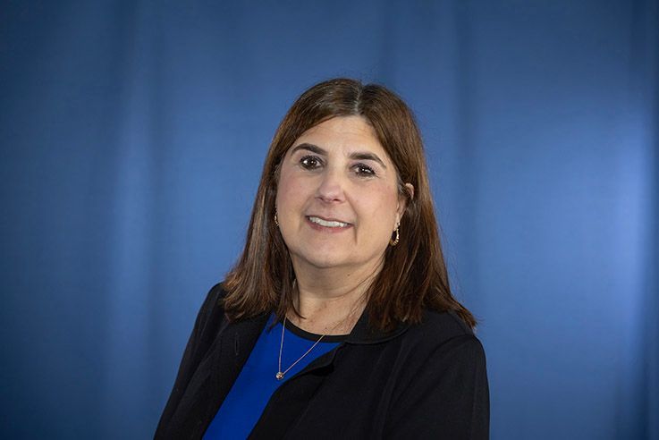Woman with brown hair, wearing a black jacket and blue shirt, smiling against a blue backdrop.