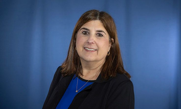 Woman with brown hair, wearing a black jacket and blue shirt, smiling against a blue backdrop.