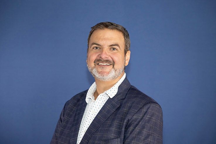 A man in a suit and white shirt is smiling in front of a blue background.