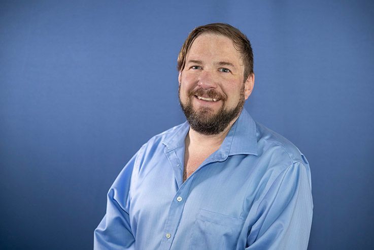 A man with a beard is wearing a blue shirt and smiling.