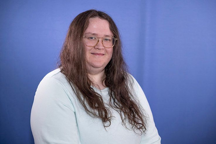A woman with long hair and glasses is standing in front of a blue wall.