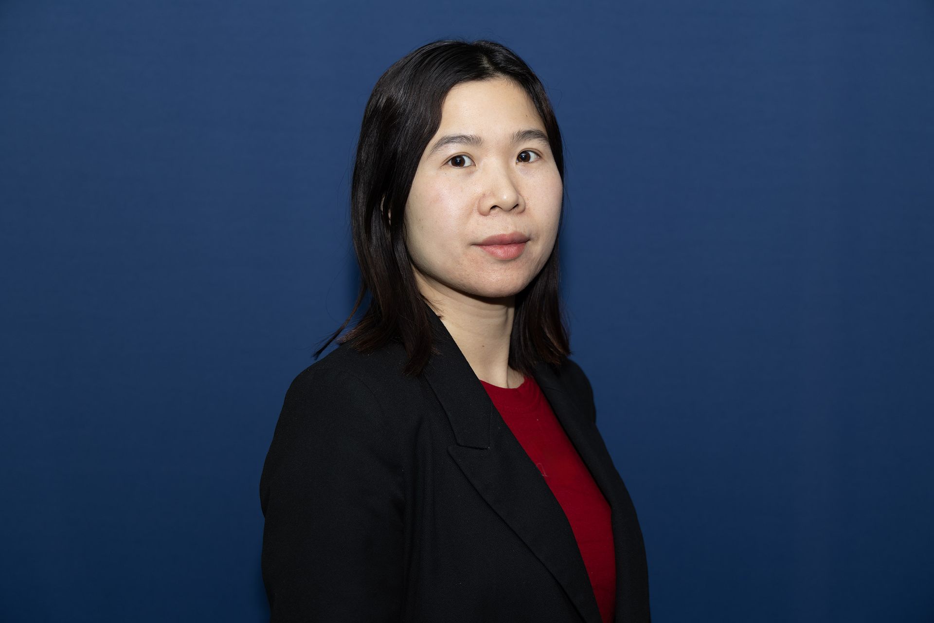 A women with dark brown hair wearing a blazer with a blue background.
