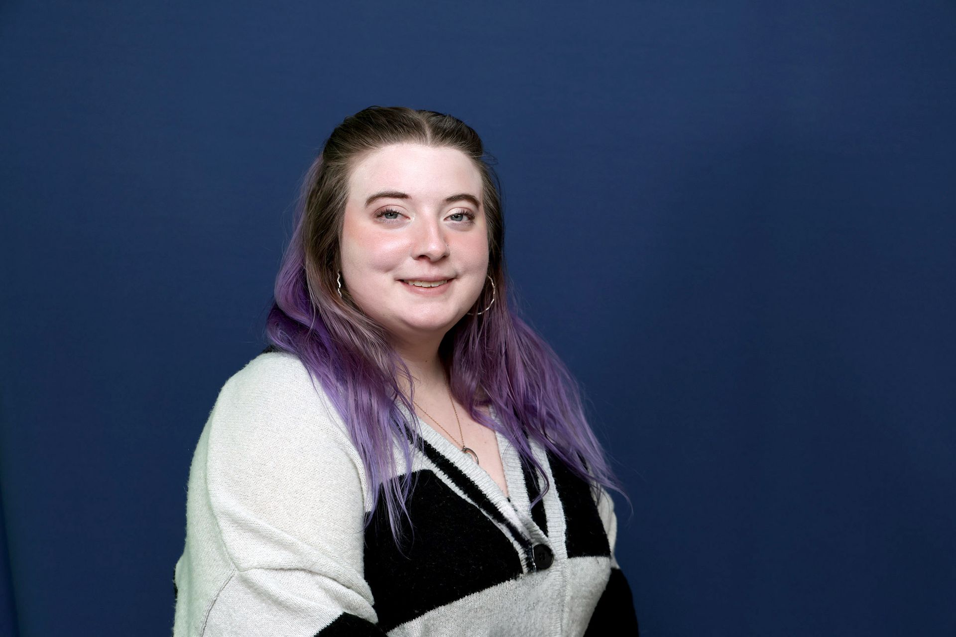 A women with brown and purple hair wearing a sweater, smiling with a blue background.