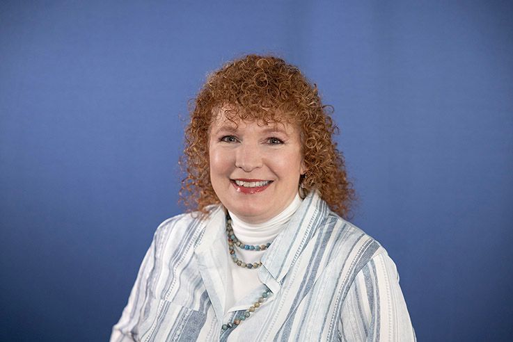Woman with red curly hair smiles wearing a white turtleneck, striped jacket, and necklace against a blue background.