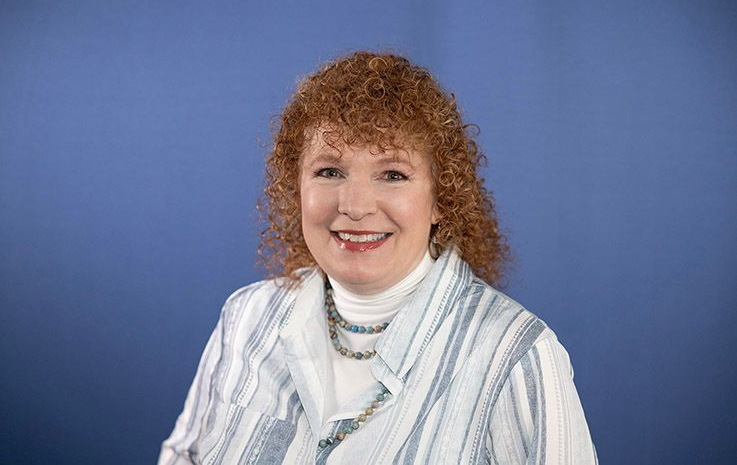 Woman with red curly hair smiles wearing a white turtleneck, striped jacket, and necklace against a blue background.