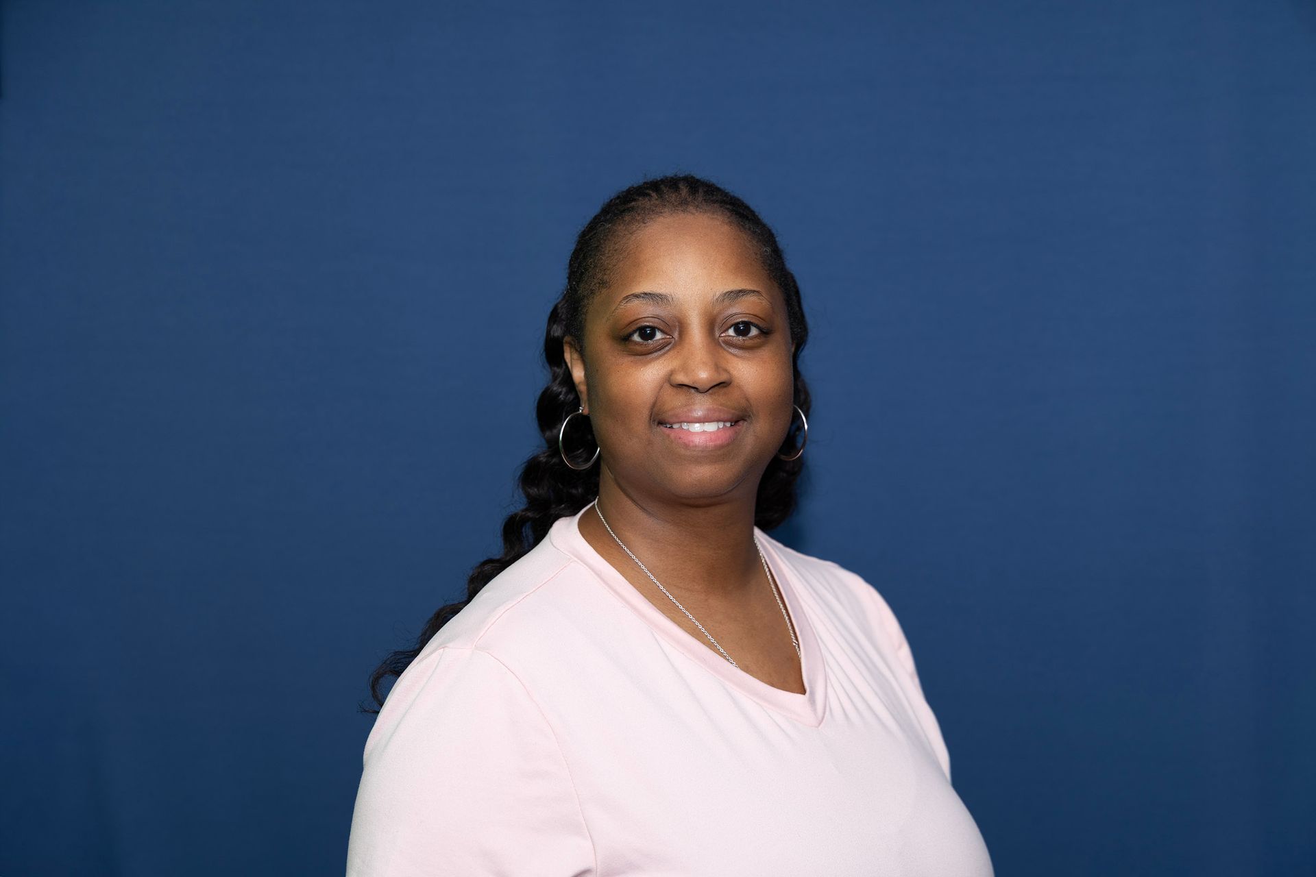 A women with long brown hair wearing a pink shirt, smiling with a blue background.