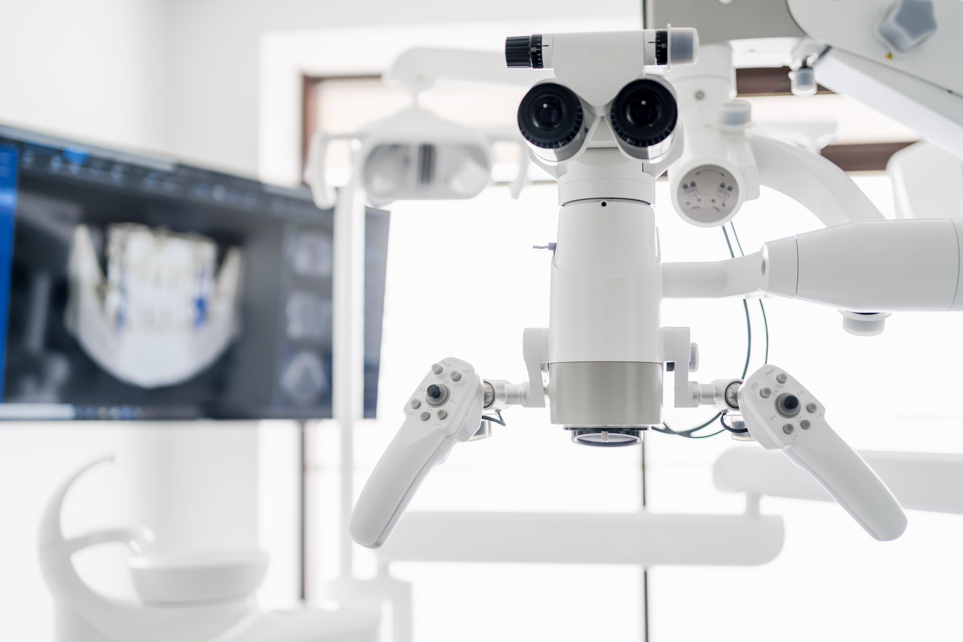 Dental microscope with attached arms, in a bright dental office; a monitor displays a dental scan.