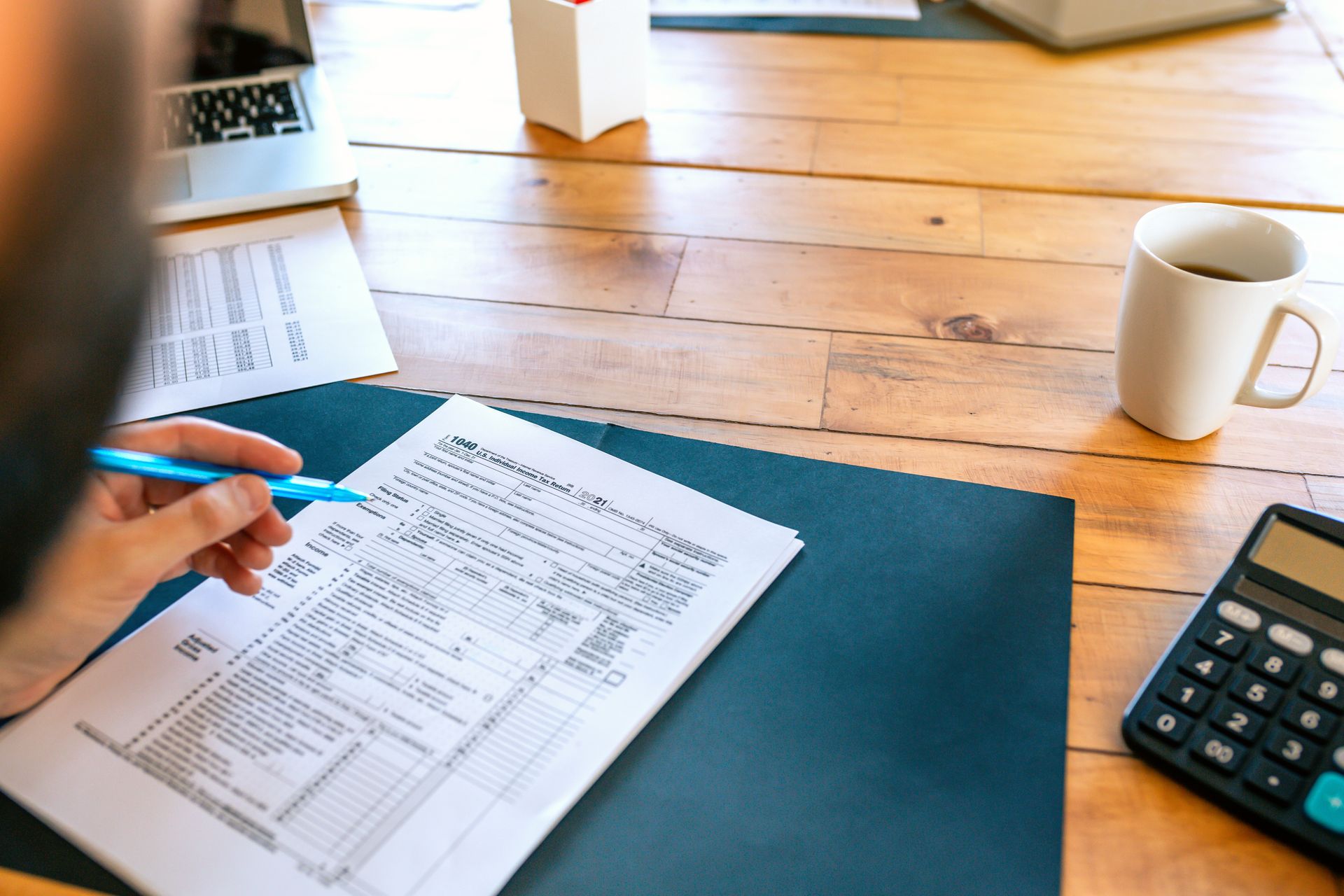 Hand with pen reviewing a printed invoice on a desk beside a laptop, coffee mug, and calculator