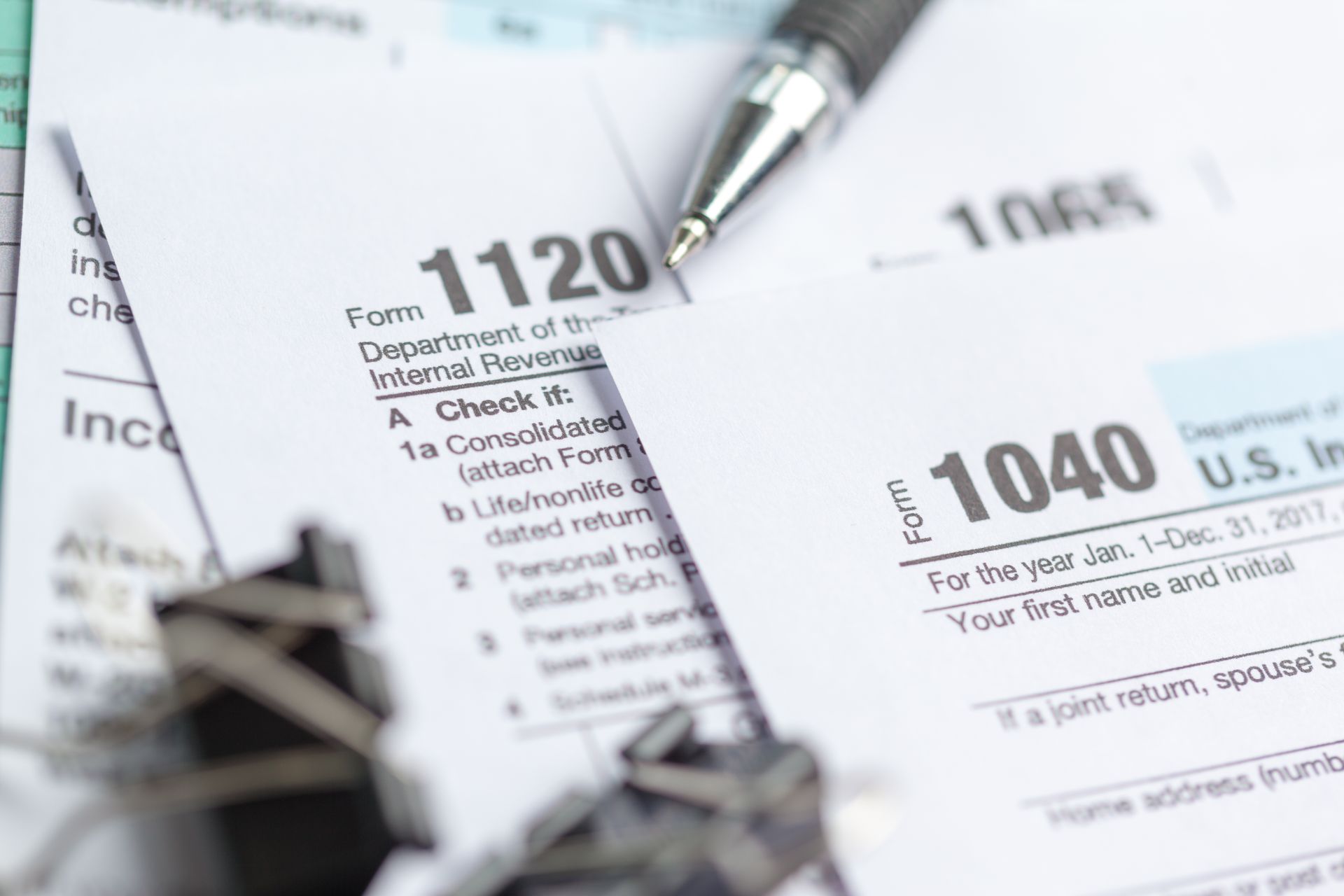 A silver pen rests on top of stacked U.S. tax forms, including IRS Forms 1120, 1040, and 1065.