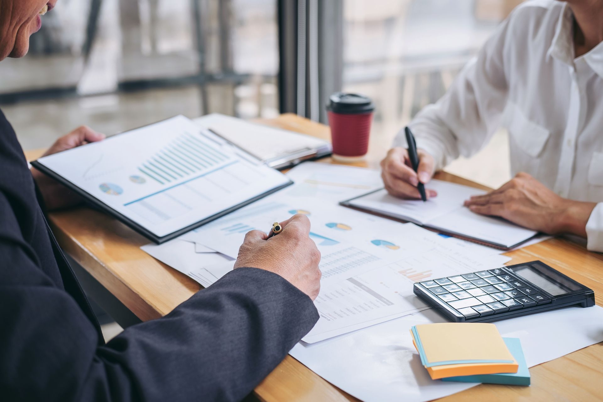 Two people in professional attire review financial charts and take notes at a desk with a calculator and coffee.