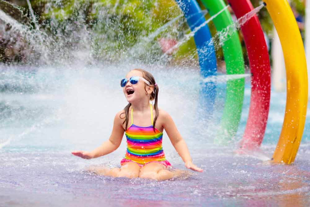 Girl In Rainbow Swimsuit And Sunglasses Enjoys Water Park Fun, Sitting Under Colorful Water Arches