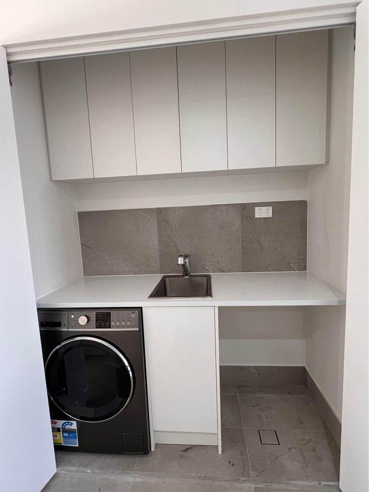 Compact laundry nook with a front-loading washer, sink, countertop, and overhead cabinets in a white alcove