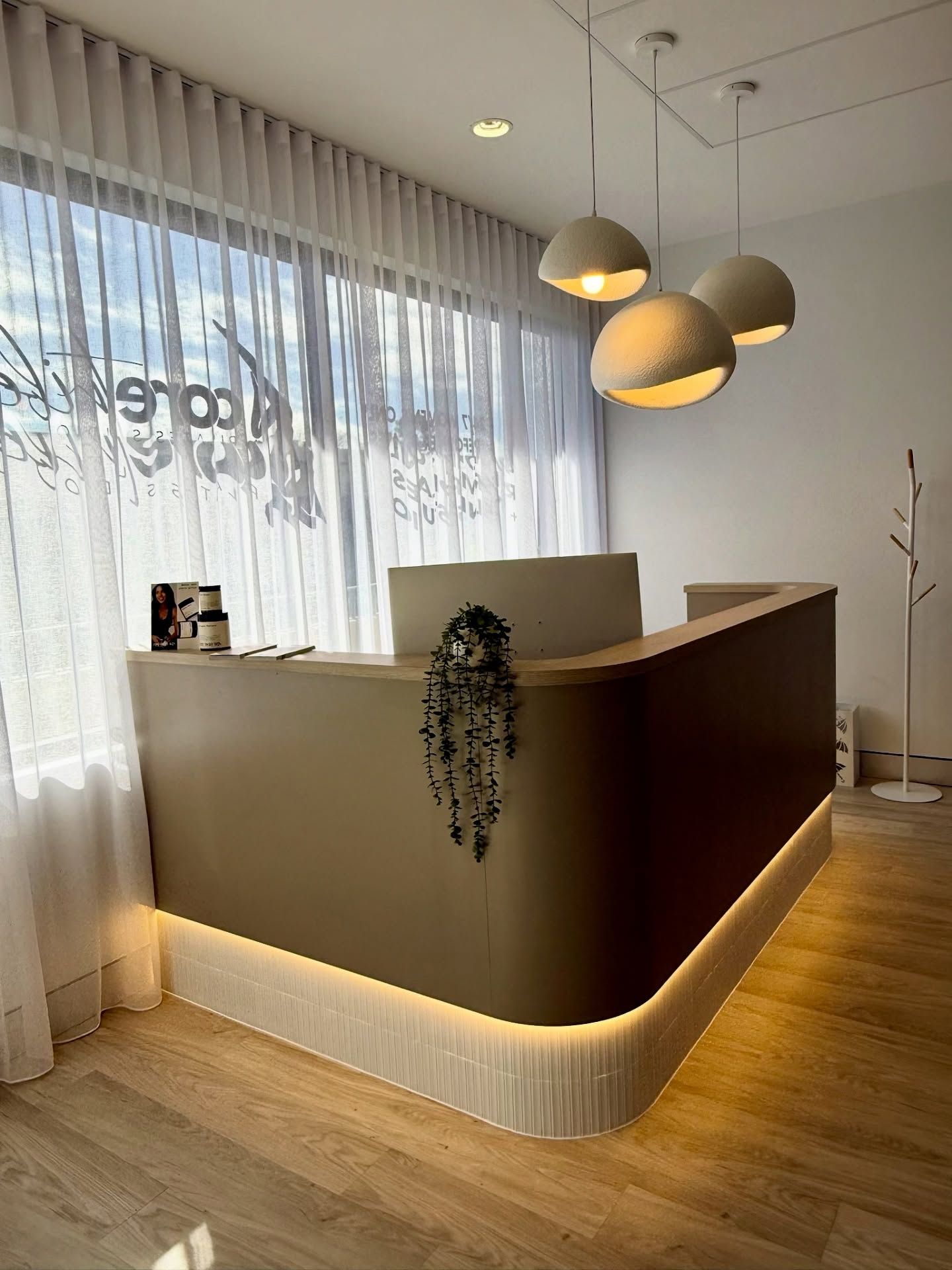 Modern reception desk with warm wood flooring, pendant lights, and sheer curtains in a bright lobby