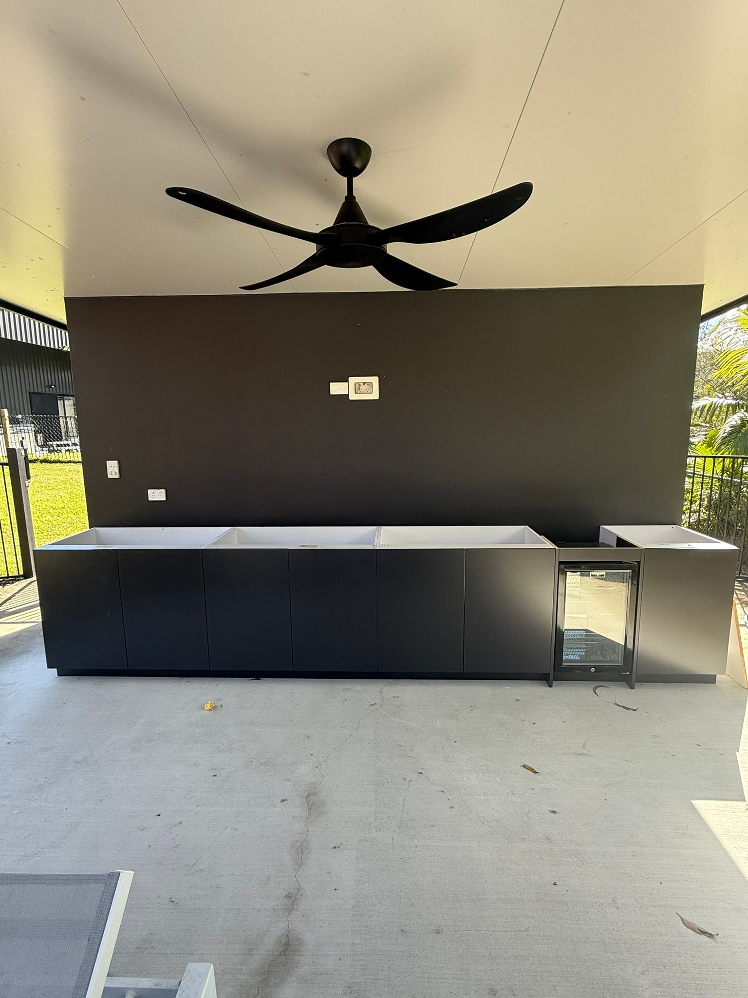 Covered outdoor patio with black ceiling fan, dark wall, long counter, and gray floor.