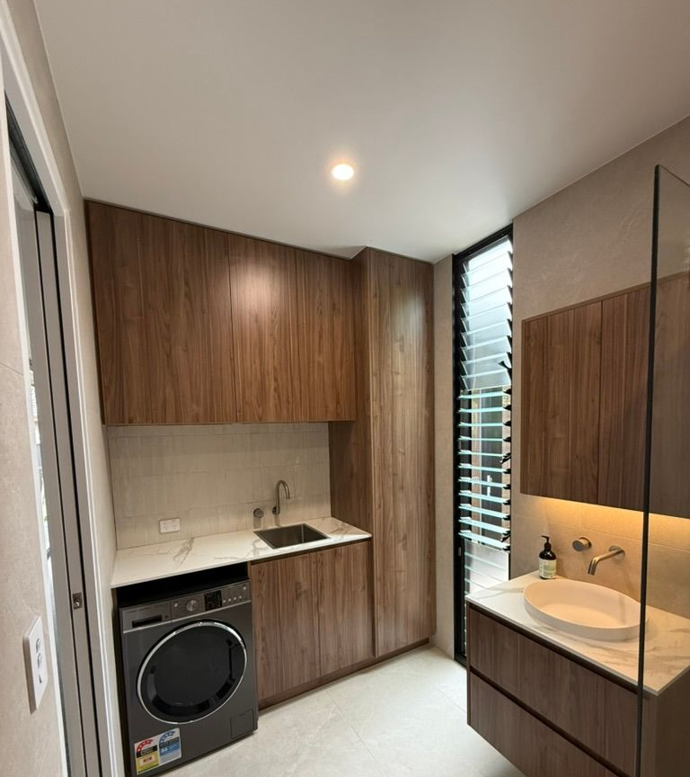 Modern laundry room with wood cabinets, stacked washer-dryer, sink, and tall narrow window