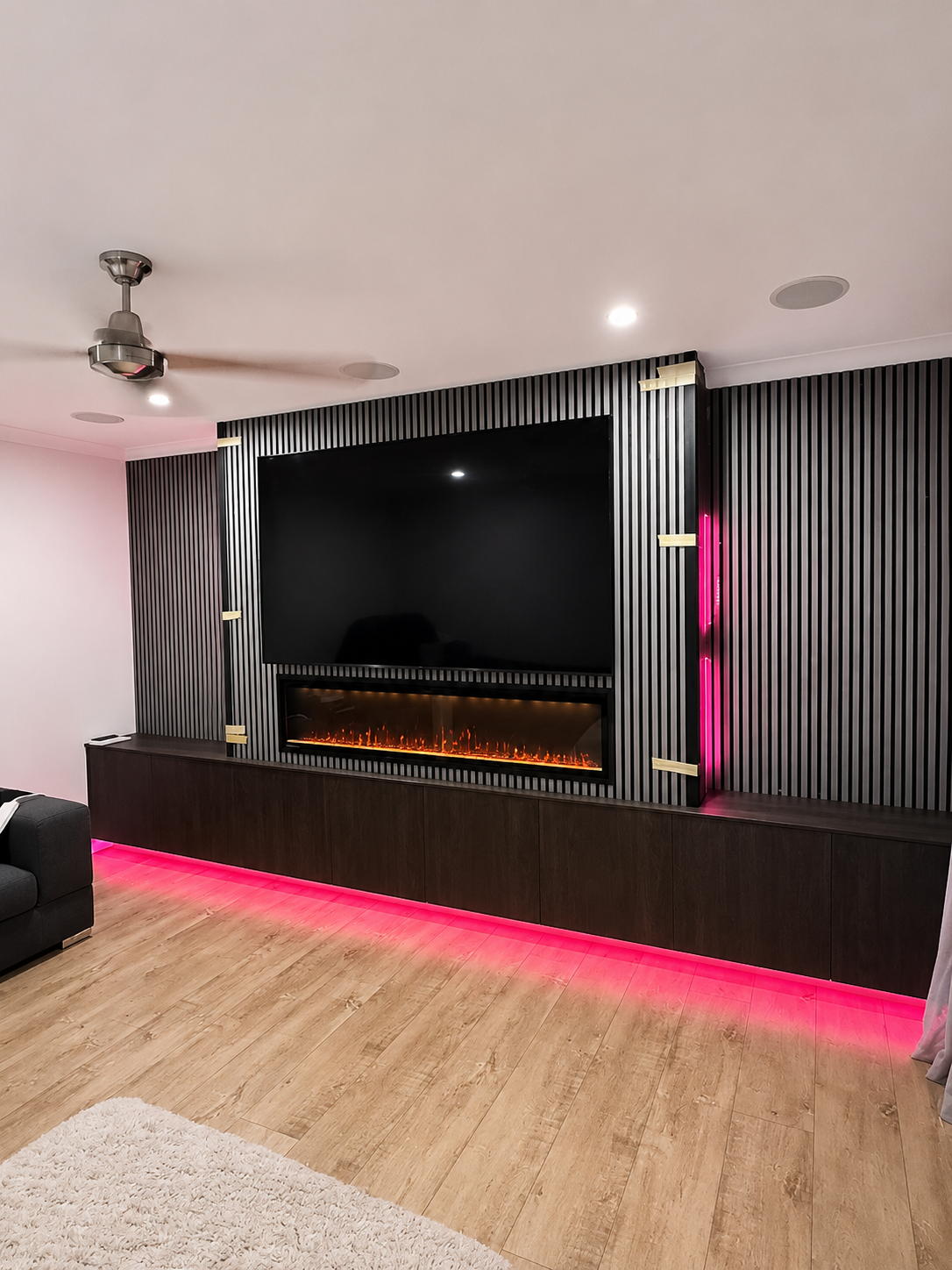 Modern living room with a large TV, electric fireplace, striped wall, pink LED lighting, and wood flooring